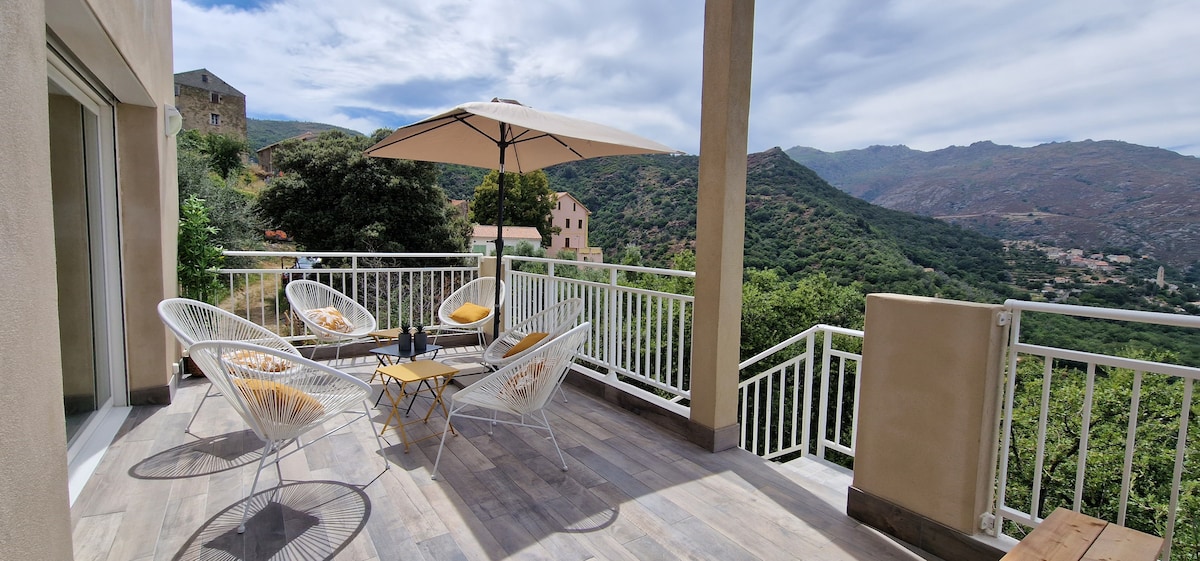 A spacious terrace features outdoor seating with two chairs and a small table, accompanied by a large umbrella providing shade. The surrounding landscape includes rolling green hills and mountains, creating a peaceful and scenic atmosphere ideal for relaxation.