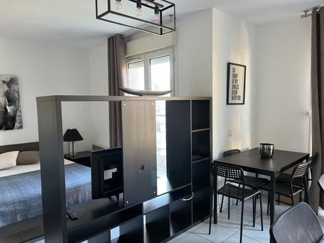 A modern apartment layout is shown, featuring a partitioned area for relaxation and dining. A spacious bed is positioned in the background, while a dining table with black chairs is visible in the foreground. Natural light enters through large windows adorned with light curtains.