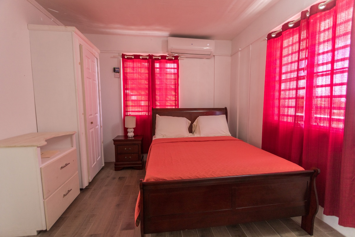A bright bedroom with red curtains complements white walls, providing ample natural light. The spacious bed is dressed in a vibrant orange coverlet, flanked by bedside tables. An air conditioning unit is installed for comfort, and a wardrobe is positioned against the wall.