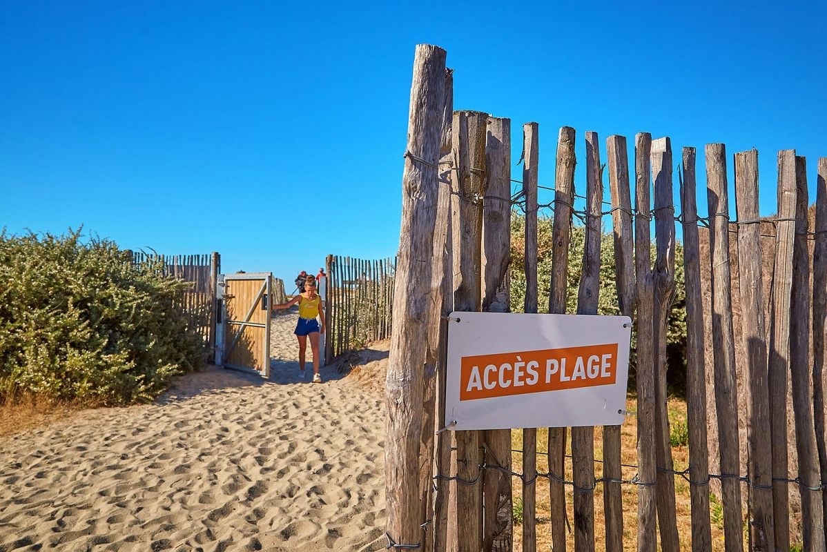 A sandy pathway leads through a wooden fence, indicating beach access with a clear sign. Lush greenery flanks the entrance, while the bright blue sky creates a serene backdrop. Individuals are visible as they walk towards the beach, enhancing the inviting atmosphere.