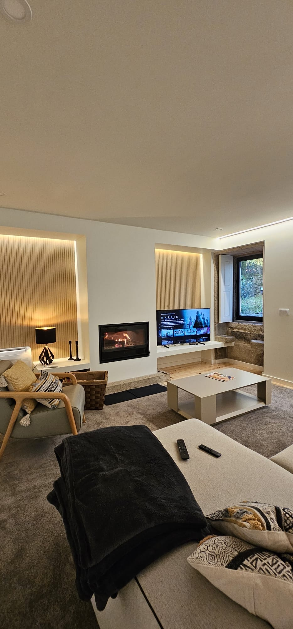 A cozy living room features a modern fireplace surrounded by light wood paneling. A flat-screen television is positioned against a stone wall, while a small coffee table is complemented by a soft couch and an armchair. Natural light enters through a window adorned with sheer curtains.