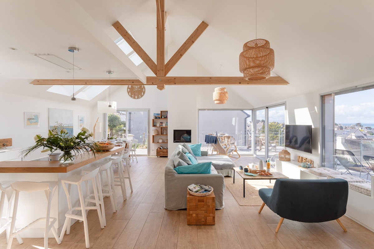 A bright and airy living room is showcased, featuring a modern design with large windows that allow natural light to fill the space. A comfortable sofa is positioned around a coffee table, while a dining area with white chairs is seen in the background, enhancing the open layout.