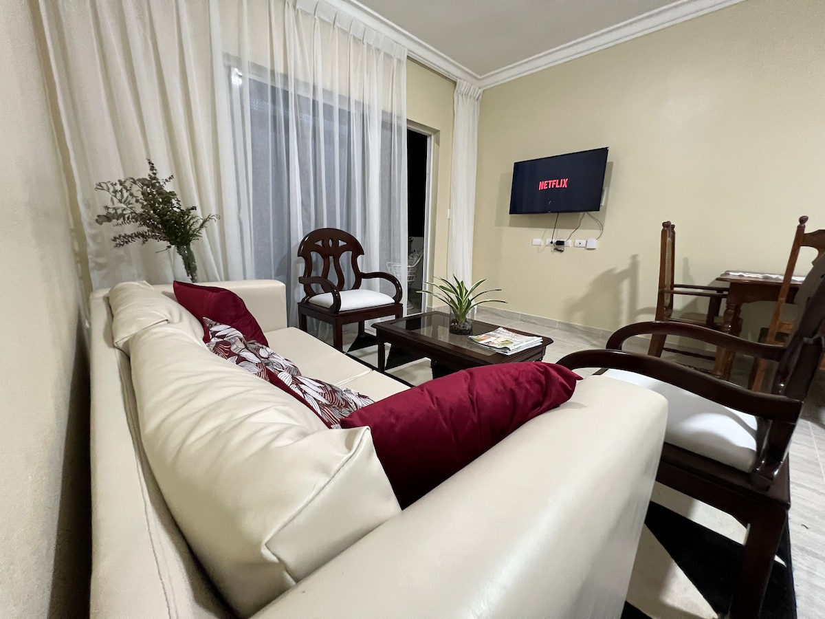 A cozy living area features a light-colored sofa adorned with decorative cushions and a glass coffee table. A television mounted on the wall displays the Netflix logo. Natural light filters through sheer curtains, illuminating the room's calming atmosphere.