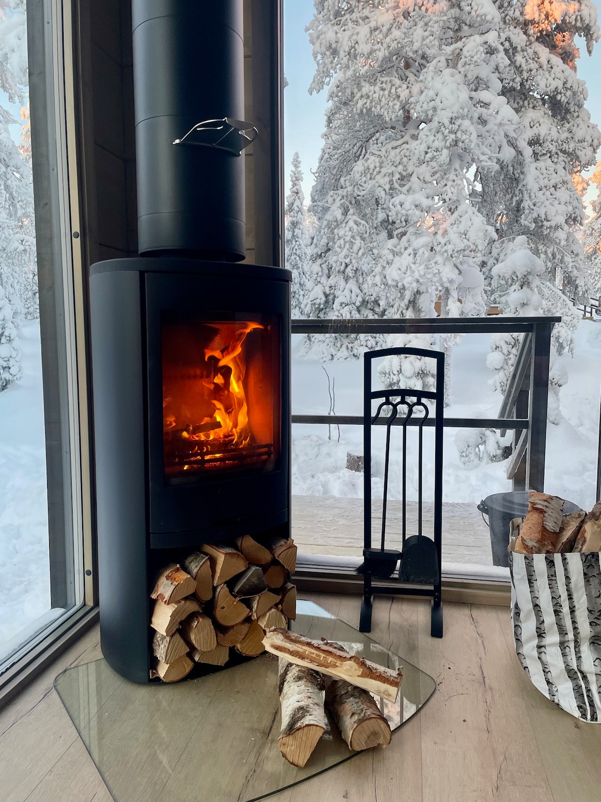 A modern black fireplace stands prominently with a glass front, displaying a warm, flickering flame. Logs are neatly stacked on a glass base, and a set of fire tools is positioned nearby. Snow-covered trees are visible through the large window, enhancing the serene ambiance.