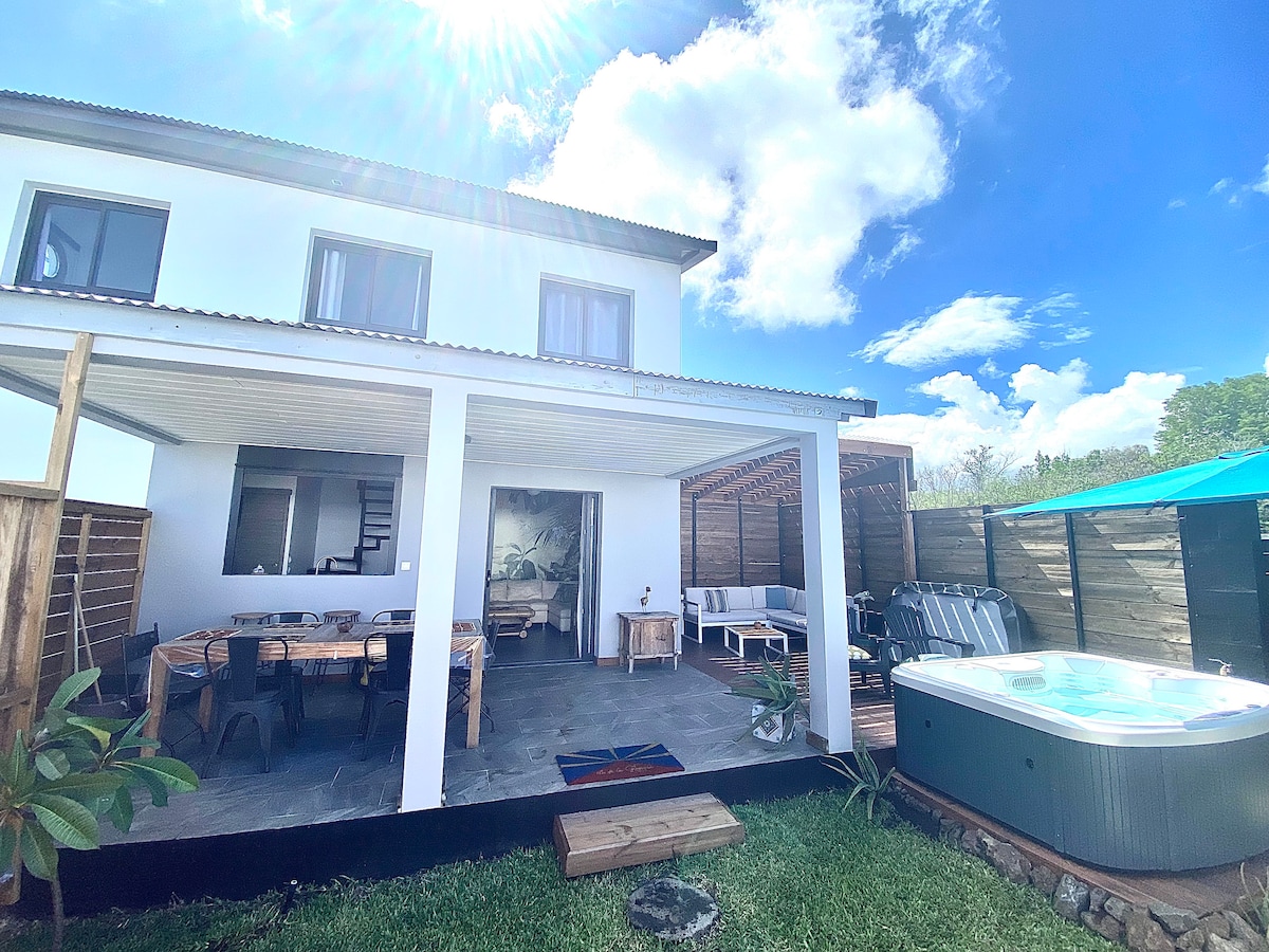 The exterior of the house features a spacious terrace with a dining area, surrounded by greenery. A high-quality spa sits nearby. The structure boasts large windows allowing natural light, complemented by a covered porch and an open design that blends indoor and outdoor living.