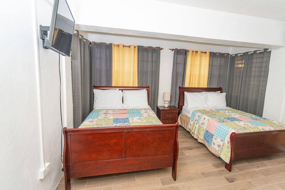 A spacious room features two wooden beds adorned with colorful patchwork quilts. Soft, natural light filters through layered curtains of gray and yellow. A small bedside table complements the beds, while a wall-mounted television is positioned for viewing.