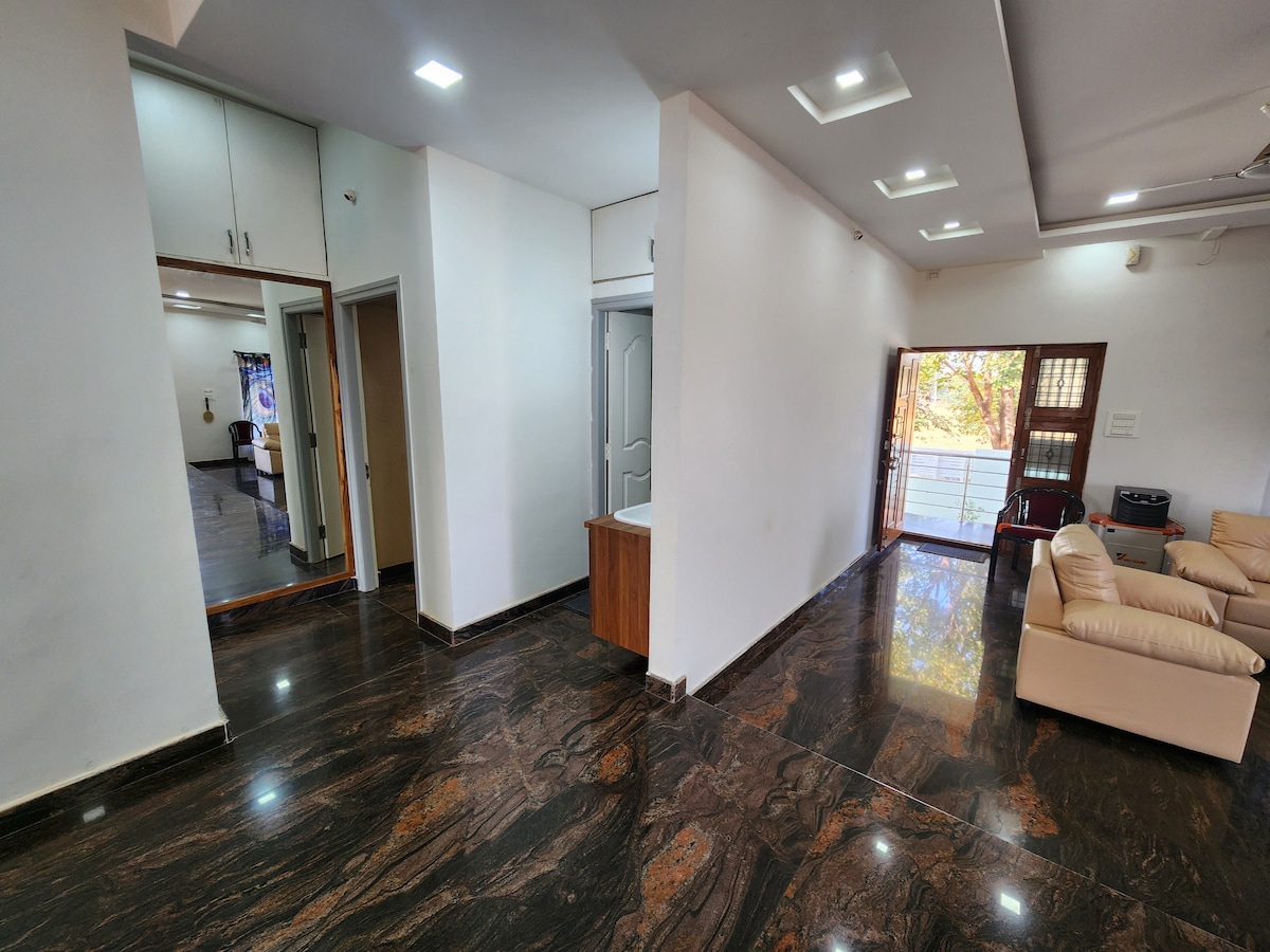 A spacious living area is showcased, featuring polished black and brown marble flooring. Natural light illuminates the room through large windows. A light-colored sofa is positioned adjacent to the entrance. Doorways lead to other rooms, enhancing the overall flow of the space.