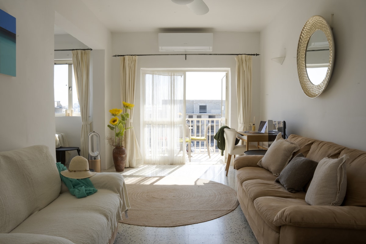 A bright living area features large sliding doors that open to a balcony, allowing natural light to fill the space. A round area rug is positioned between a two-tone couch and a chair. Decorative plants and a large mirror enhance the welcoming atmosphere.