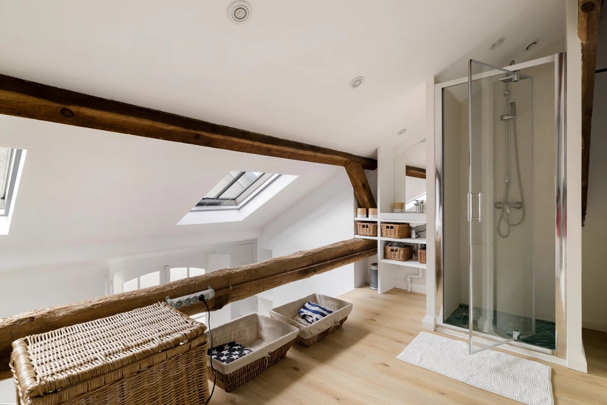 An open mezzanine space features exposed wooden beams contrasting with white walls. Natural light enters through skylights. A modern shower with transparent glass doors is visible, alongside storage baskets and a wicker basket, creating an organized, functional area.
