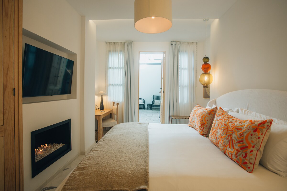 A cozy sleeping area features a spacious bed adorned with colorful decorative pillows. An electric fireplace is positioned below a wall-mounted TV. Soft light enters through sheer curtains, illuminating the minimalist decor that creates a serene ambiance.