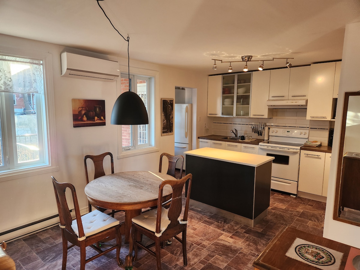 A functional kitchen and dining area features an island with seating, surrounded by a round dining table and four wooden chairs. Natural light streams through the windows, illuminating the modern cabinetry and appliances, including a stove and refrigerator.