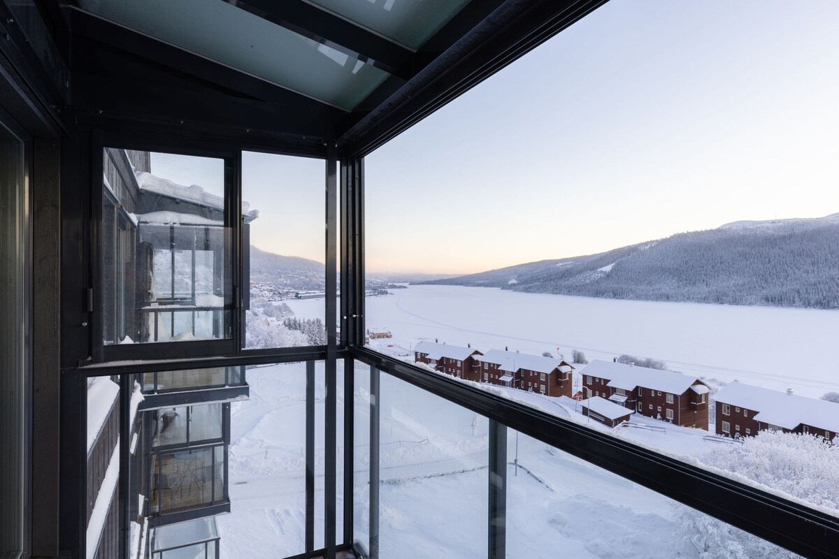 A panoramic view is presented from a glass-enclosed balcony, revealing a snow-covered landscape. The frozen lake stretches into the distance, framed by forested hills. Nearby buildings are partially visible, contributing to the serene winter scenery under soft, early morning light.