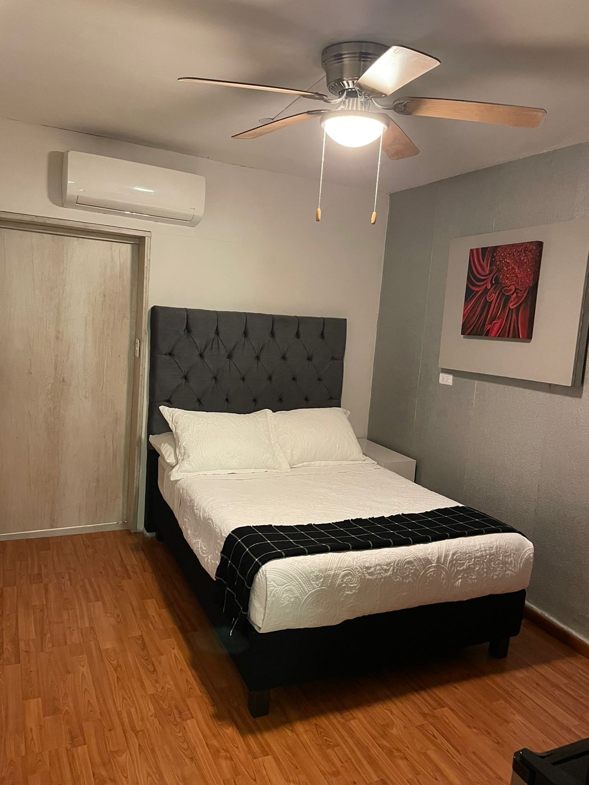 A comfortable sleeping area features a queen-sized bed with a tufted headboard, complemented by light bedding and a dark throw. A ceiling fan circulates air above, while a wall-mounted air conditioning unit provides climate control. A piece of wall art and a light wood door add depth to the space.