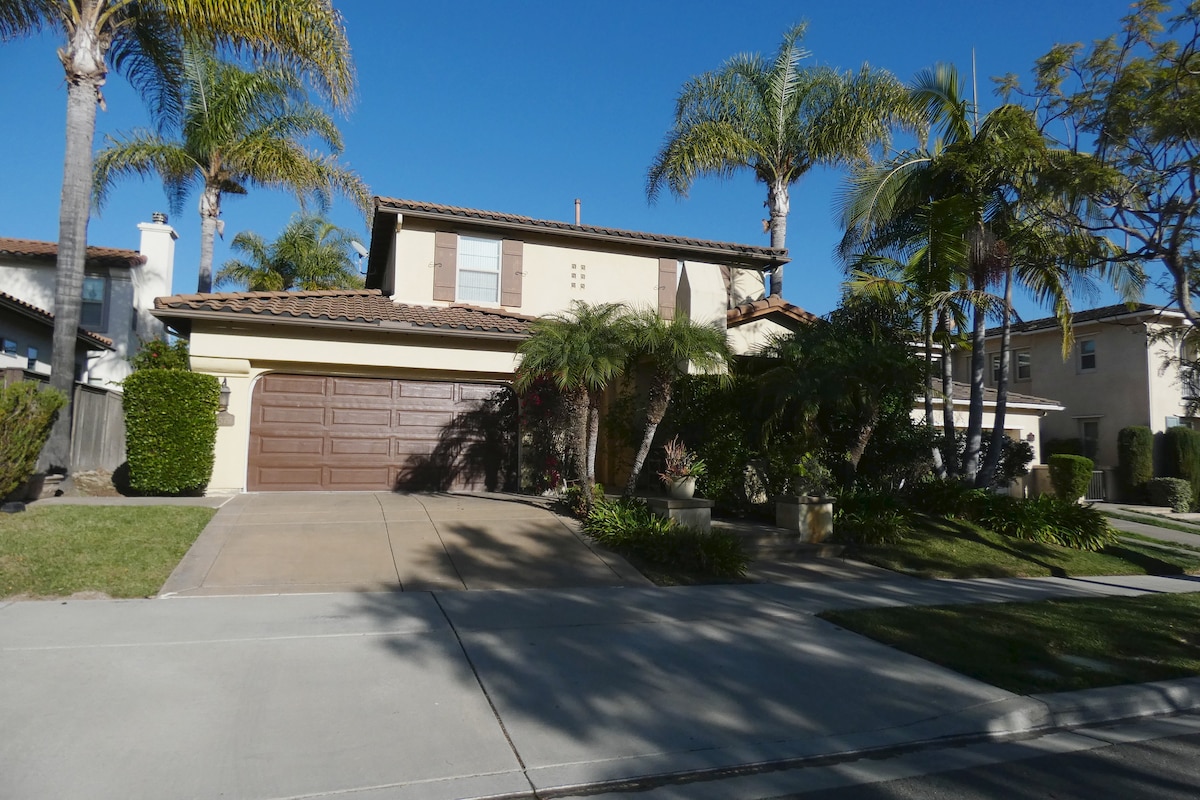 A two-story house is framed by tall palm trees and landscaped greenery. A spacious driveway leads up to a brown garage door, with a mix of stone and stucco exterior visible. The clear blue sky provides a bright backdrop.