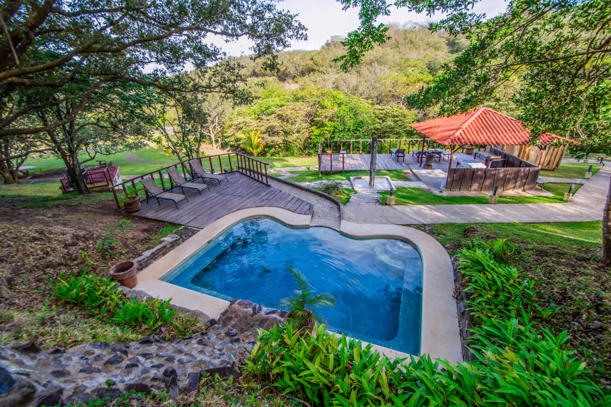 A heart-shaped pool is framed by lush greenery, with wooden decking surrounding it for relaxation. A covered pavilion with a red roof is visible in the background, providing shaded seating areas. The landscape features gentle hills and a variety of tropical plants.