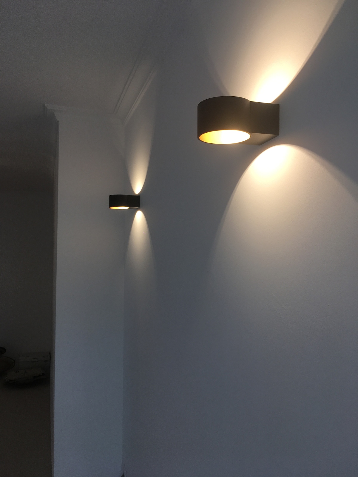 Two modern wall sconces are positioned on a white wall, illuminating the space with warm light. The light casts soft shadows, enhancing the simplicity of the decor.