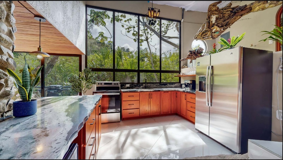 A spacious kitchen features wooden cabinetry and marble countertops, complemented by a sleek stainless-steel refrigerator. Natural light floods the area through large windows, showcasing views of the surrounding greenery. Potted plants add a touch of nature to the modern design.