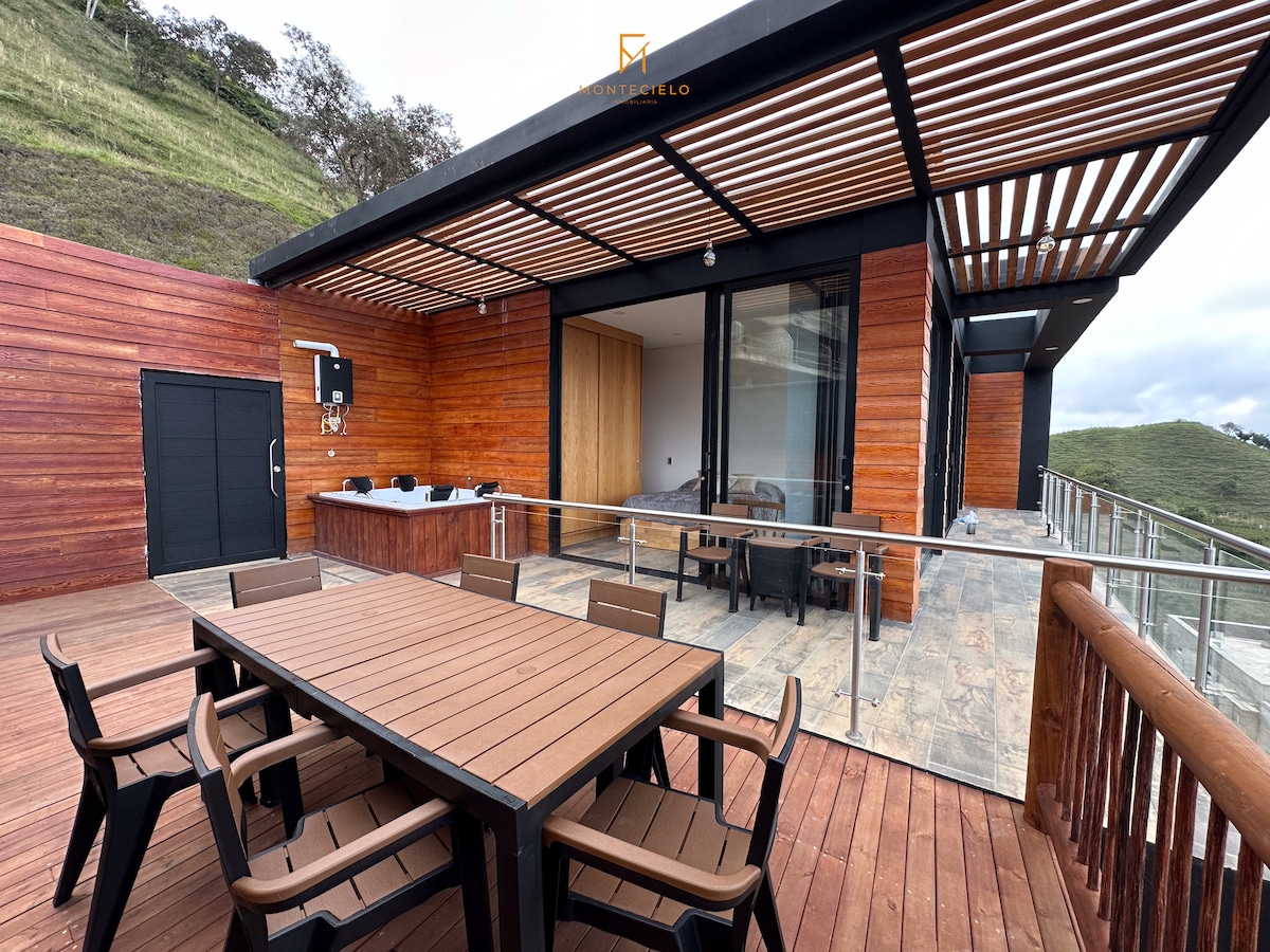 An outdoor terrace features a large wooden dining table surrounded by several chairs. The space highlights a sleek wooden exterior with large glass doors that offer views of the landscape. A built-in grill is visible against the wall.