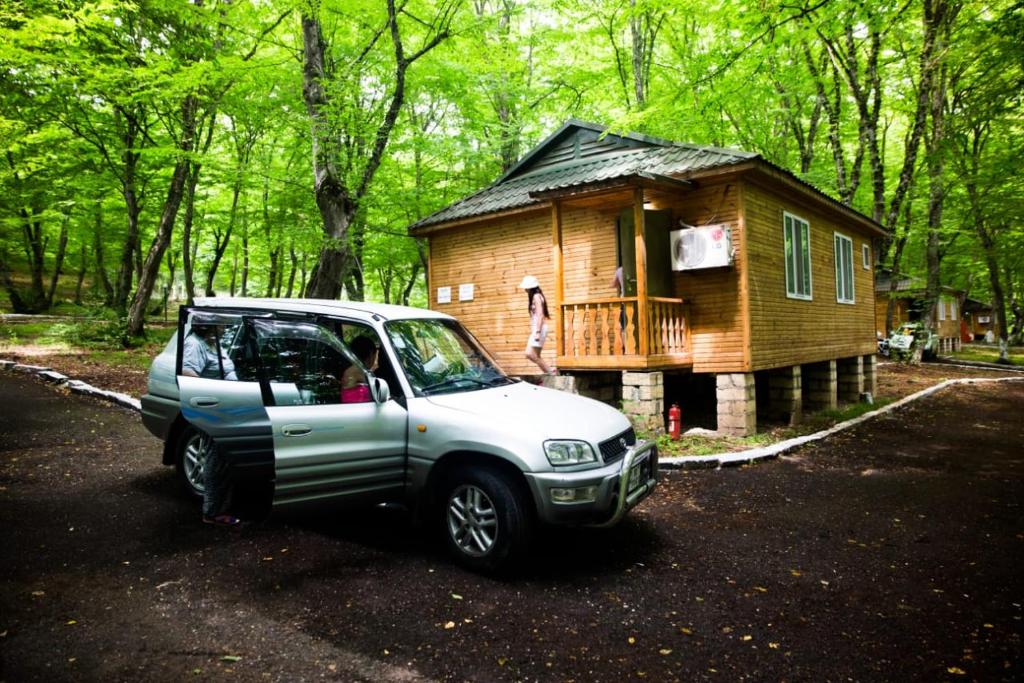 A wooden cottage is situated amidst a lush green forest. A silver SUV is parked in the foreground, with a pathway leading toward the cottage's entrance. The area is well-defined by trees, providing a natural and serene backdrop.