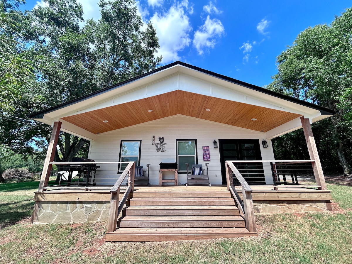 A welcoming exterior of a single-story retreat is shown, featuring a broad porch with wooden steps. Comfortable seating areas are arranged on the porch, with a gently sloping roof and decorative elements. Surrounding lush trees provide shade and enhance the outdoor space.