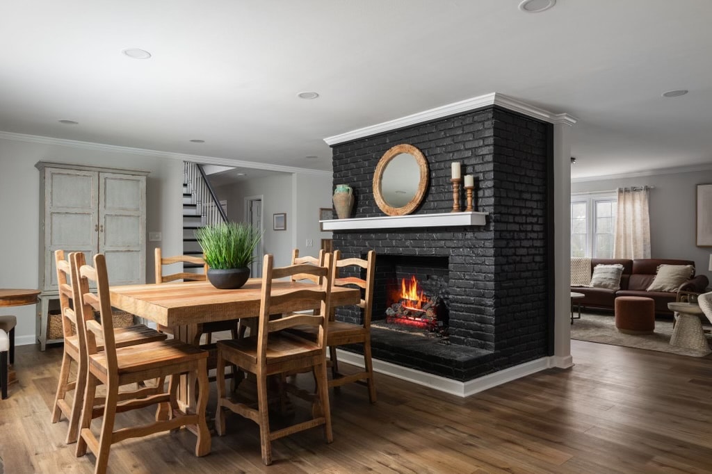 A wooden dining table with six chairs is positioned near a black-brick fireplace, which features a round mirror above it. A potted plant sits on the table, and a cozy living area is visible in the background, complemented by natural light from large windows.