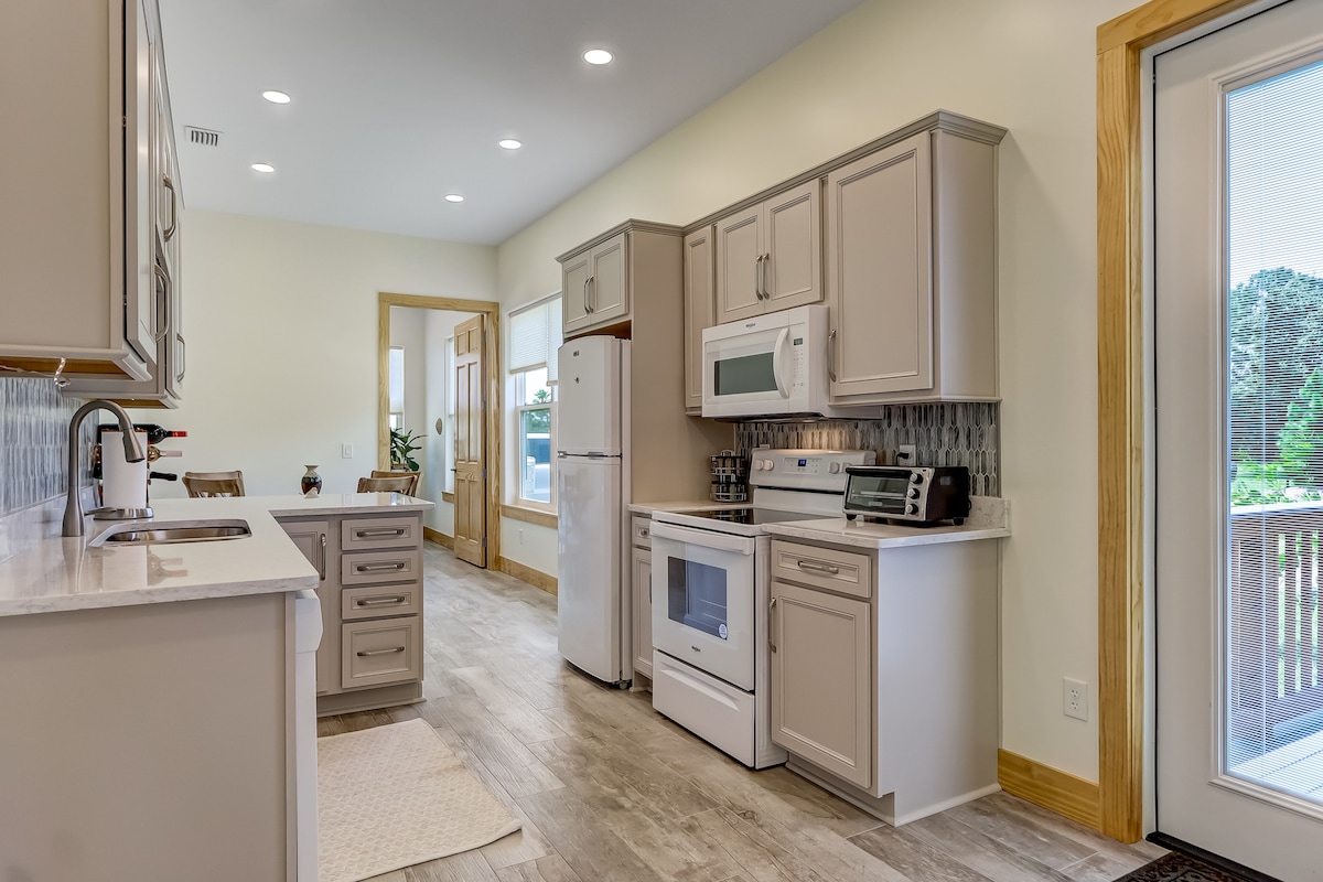 A well-equipped kitchen features light-colored cabinets and modern appliances, including a refrigerator and microwave. Natural light brightens the space from sizeable windows. A small dining area with simple chairs is visible, accompanied by a sliding door leading to an outdoor deck.