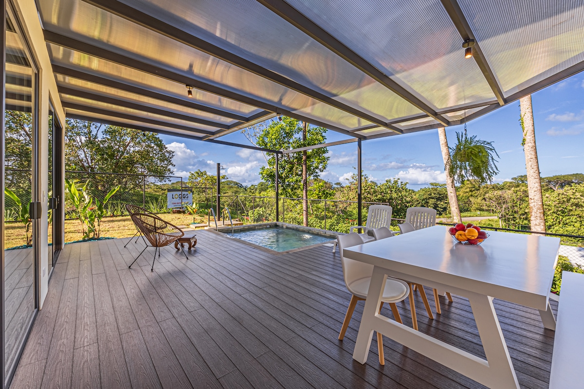 A modern outdoor dining space features a table with six chairs, complemented by a relaxing lounge chair. A private pool is visible in the background, surrounded by lush greenery. The area is covered with a sleek awning, creating a comfortable atmosphere for gatherings.