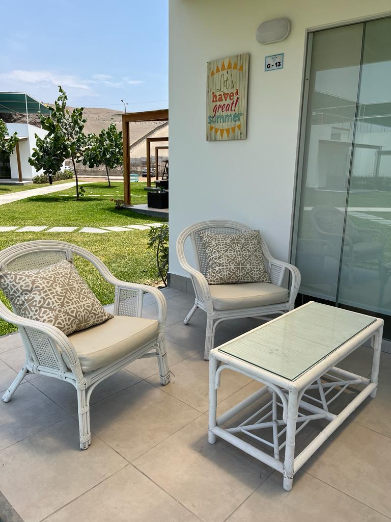 A cozy seating area is presented outdoors, featuring two comfortable white wicker chairs with cushions and a glass-topped table. The background showcases green grass and trees, enhancing the inviting space. A cheerful sign displaying a summer phrase adds a touch of charm.