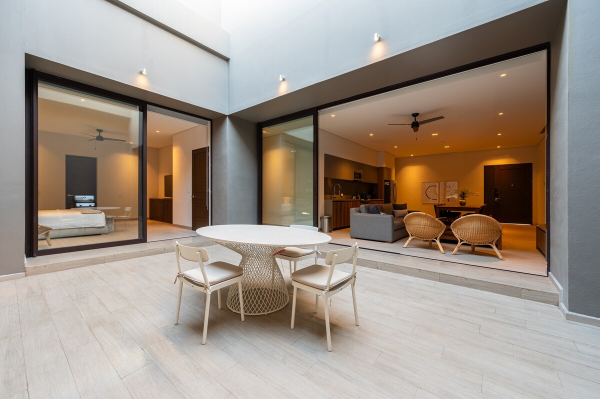 An inviting courtyard is presented, featuring a round dining table surrounded by four light-colored chairs. Large glass doors frame the living and sleeping areas, allowing natural light to fill the space. The elegant floor tiles contribute to a modern aesthetic.