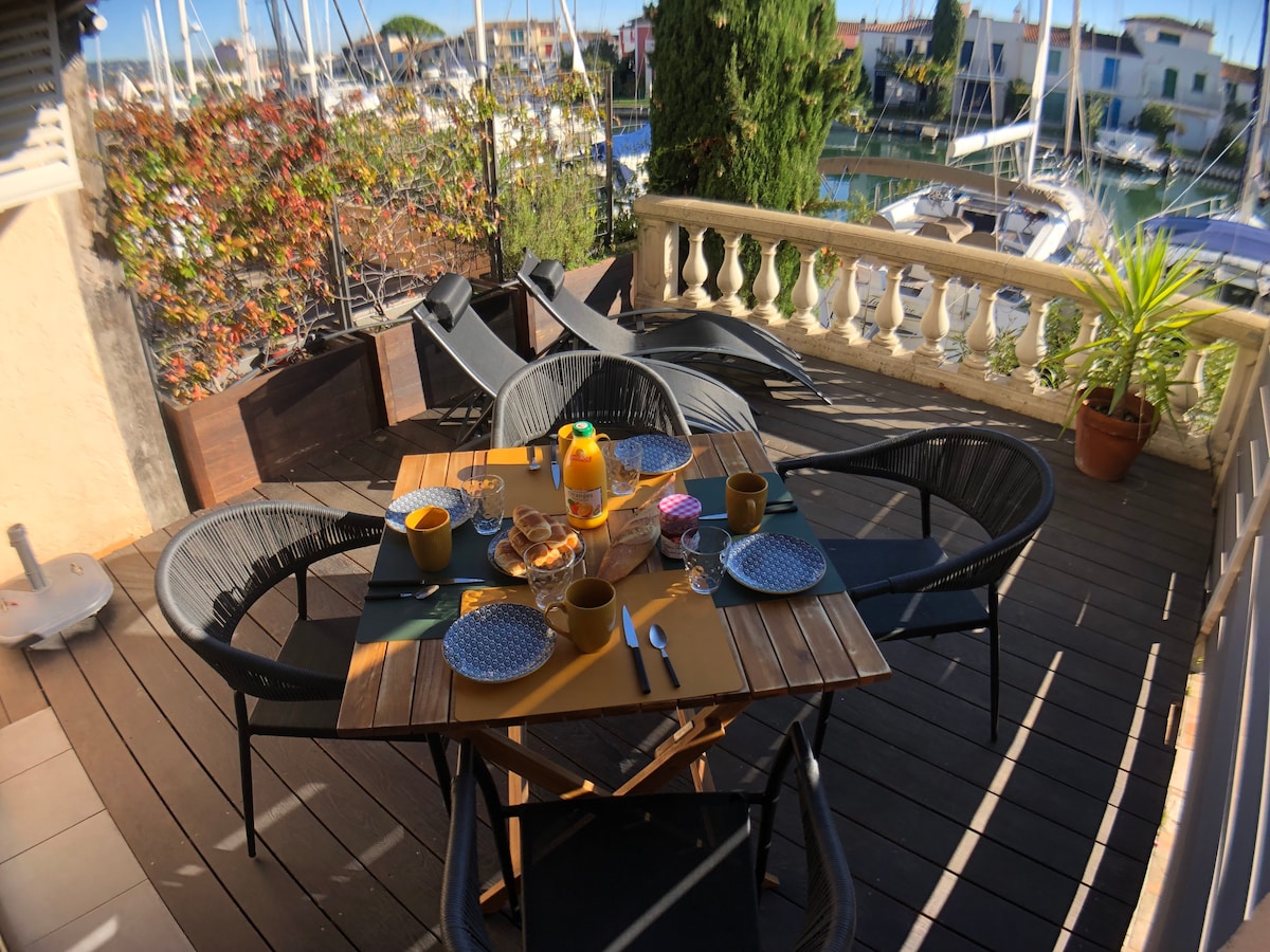 An outdoor terrace is featured, furnished with a wooden table surrounded by six black chairs. Set on the table are colorful plates, cutlery, and drinks. The background displays boats in the water, with greenery and decorative elements adding a natural touch.