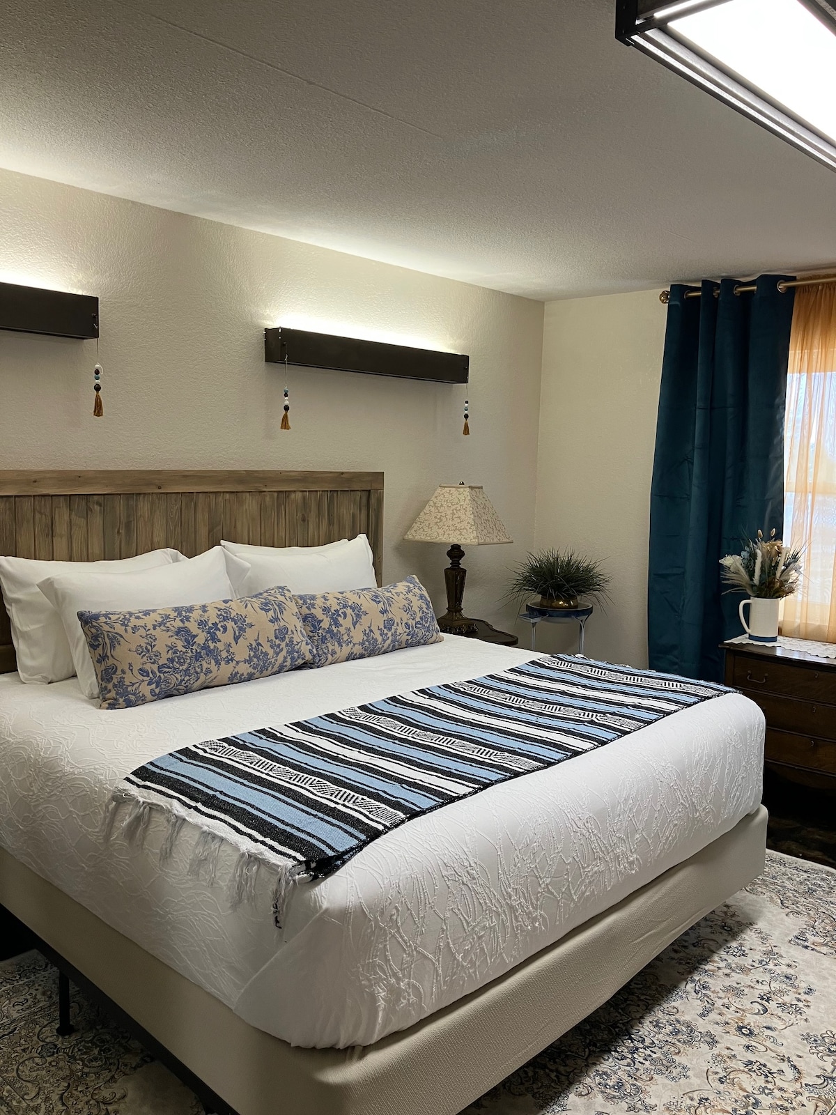 A well-appointed bedroom features a spacious bed dressed in crisp white linens and accented by a decorative blue and white throw. Soft lighting from wall sconces complements the natural light filtering through sheer curtains. A bedside lamp and greenery add warmth to the space.