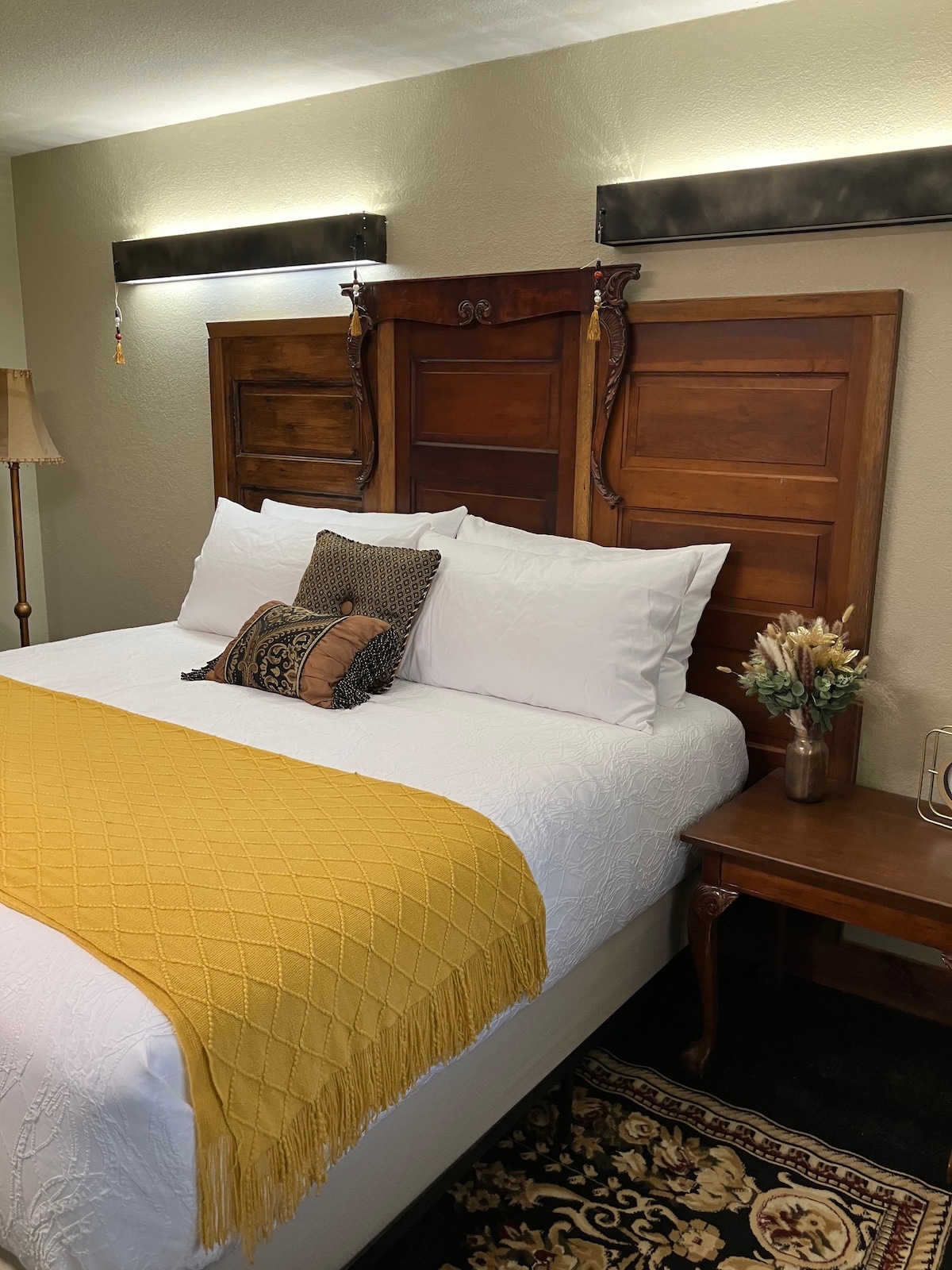 A spacious bed is framed by an elegant wooden headboard, complemented by decorative pillows. A warm yellow blanket adds a touch of color, while a side table holds a small vase with dried flowers. Soft lighting enhances the inviting atmosphere.