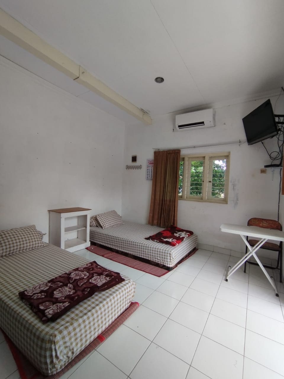 The room features two beds with checkered bedding positioned on red mats. A small wooden cabinet is placed beside the beds. An ironing board is set up in the corner, and a wall-mounted television is visible. Natural light enters through a window with curtains.