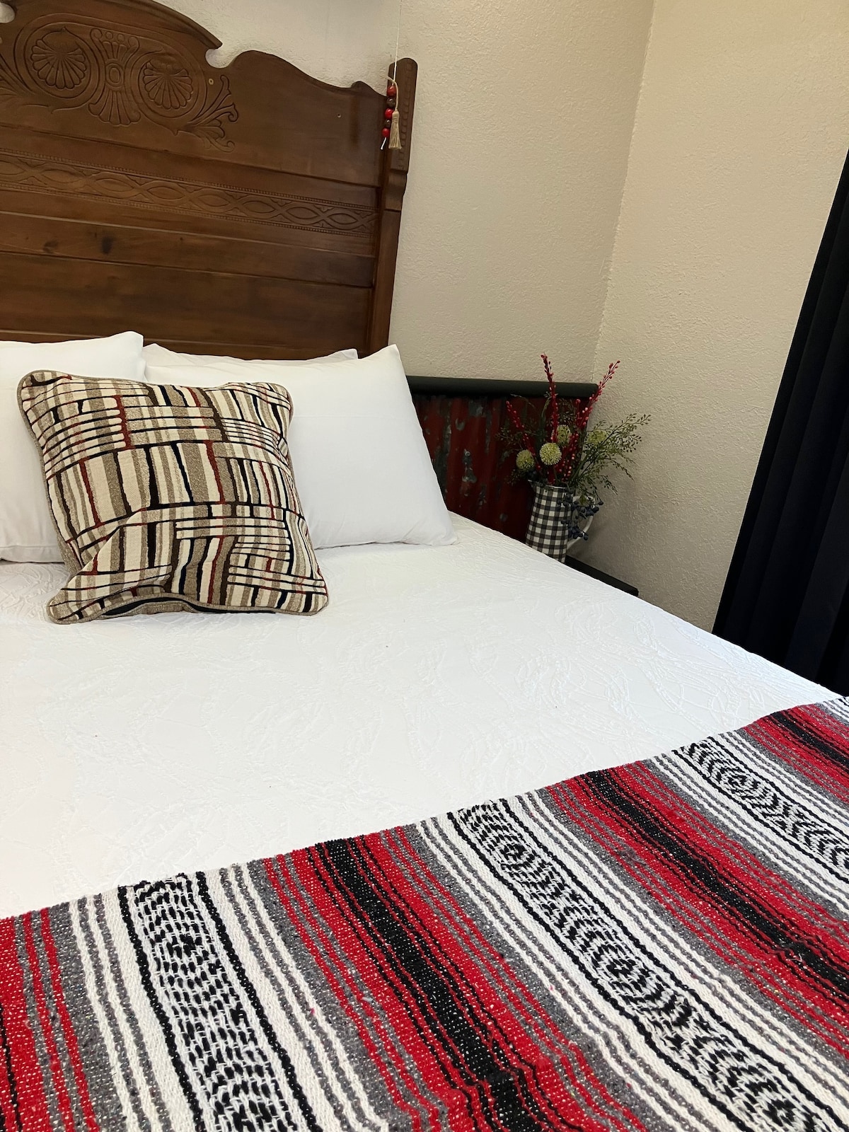 A neatly made bed features a white quilt and a decorative pillow with a plaid pattern. A colorful woven blanket in red and black drapes across the foot of the bed. A small vase with foliage accents the corner of the room.