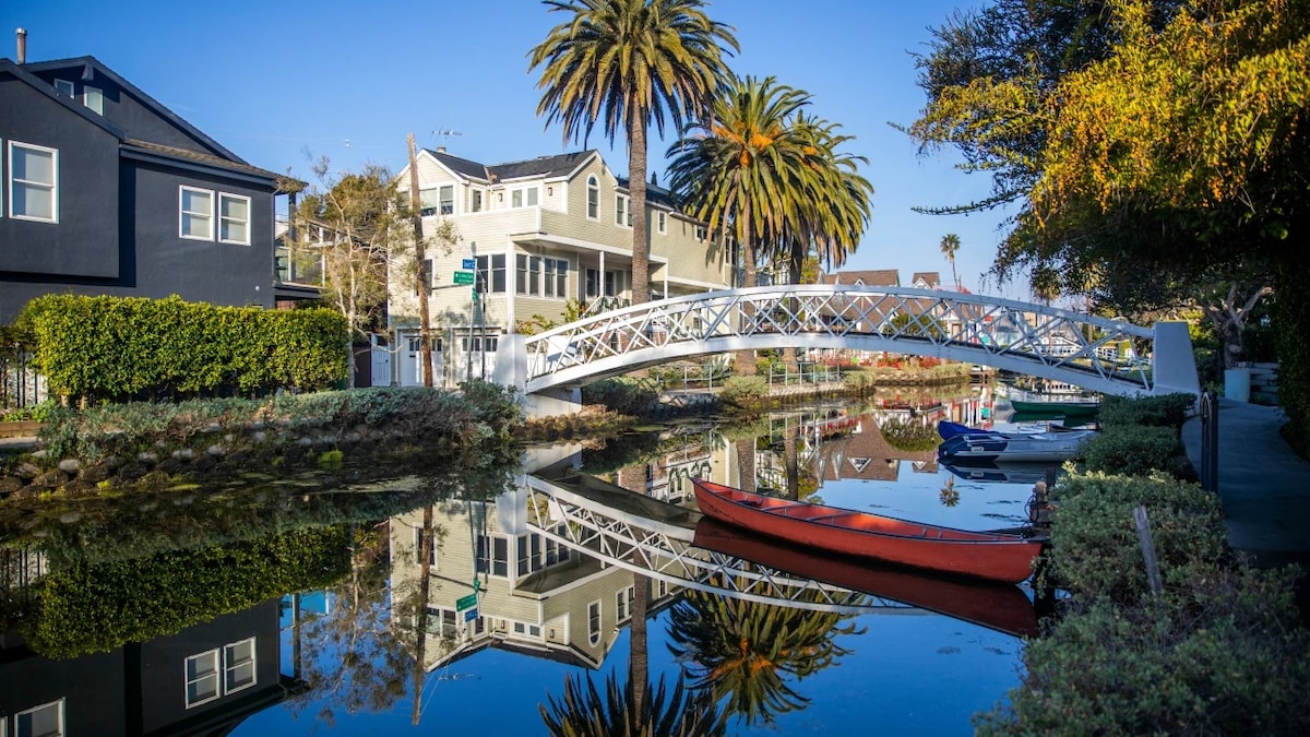 A serene view of calm waters reflects palm trees and charming houses lining the canal. A white footbridge crosses the water, leading to lush greenery, while a red kayak rests along the shoreline, adding a pop of color to the tranquil scene.