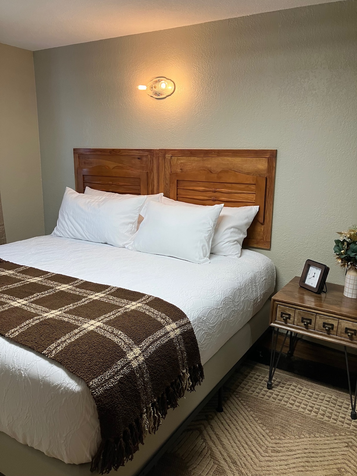 A cozy bedroom is arranged with a spacious bed featuring two headboards. Two white pillows and a brown plaid throw blanket are neatly placed on top. A wooden nightstand with a small lamp and decorative items sits beside the bed, while textured flooring adds warmth to the space.