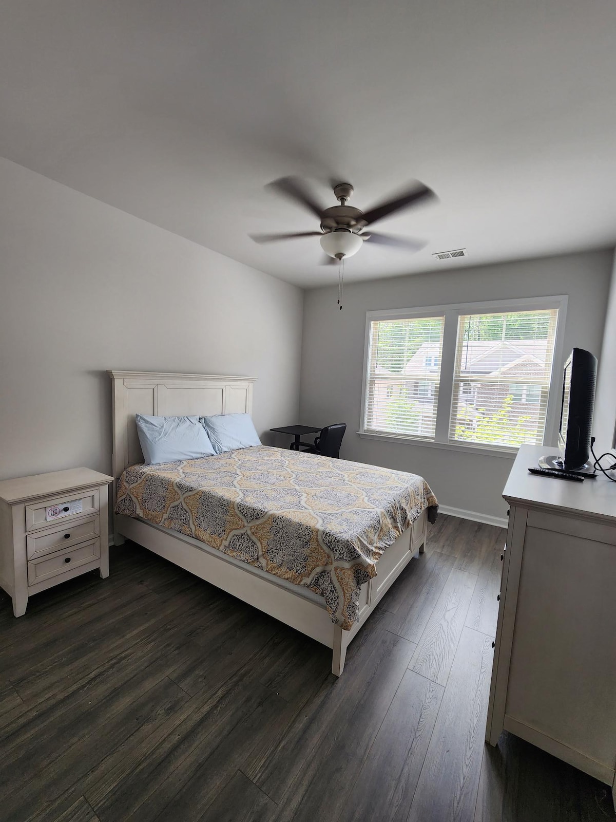 A well-lit bedroom featuring a queen-size bed with a patterned comforter. A nightstand holds a small lamp. A flat-screen TV sits on a dresser, and a chair is positioned near the window, which allows natural light to enter the space.