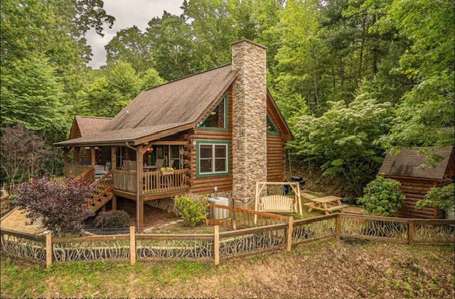 The cabin features a rustic design with natural wood accents and a stone chimney. A welcoming porch is adorned with sturdy railings, leading to a landscaped yard filled with trees and colorful shrubs. A separate structure is visible nearby, enhancing the private setting.