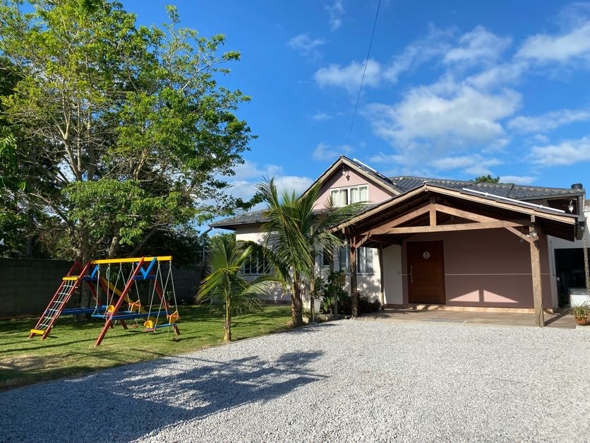 A welcoming house features a gravel driveway and is surrounded by greenery. A spacious front porch with a wooden roof offers shade. To the left, a colorful swing set is situated on the lawn, providing a playful space for children.