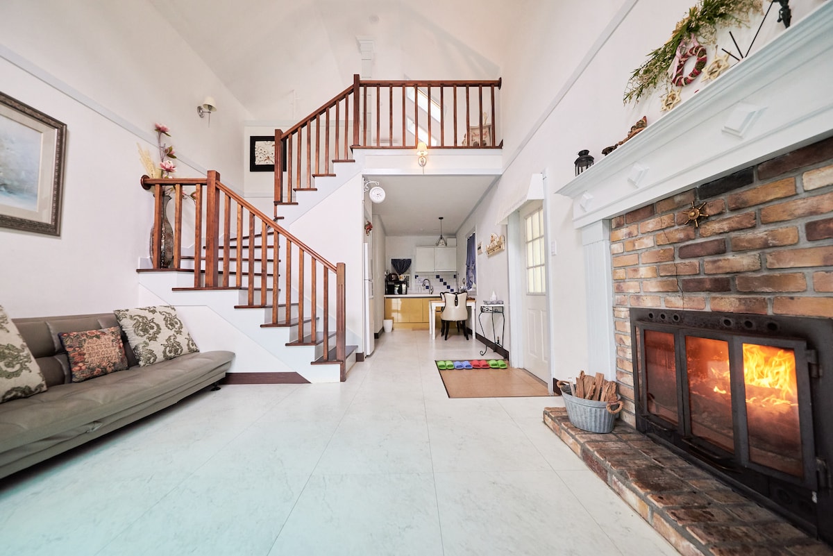 A spacious living area features a sofa with decorative pillows, complemented by a wooden staircase leading to an upper level. A traditional fireplace, set against a brick wall, adds warmth to the space. Natural light flows through the nearby windows, illuminating the open layout.