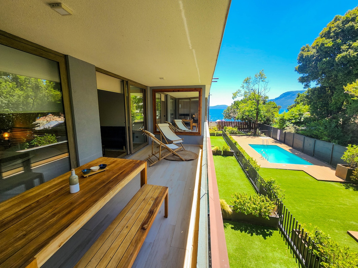 A spacious balcony is visible, featuring a wooden dining table and two lounge chairs. Below, a well-kept garden includes a clear pool surrounded by manicured grass and a wooden deck. A scenic view of the lake and trees is framed by the balcony's glass doors.