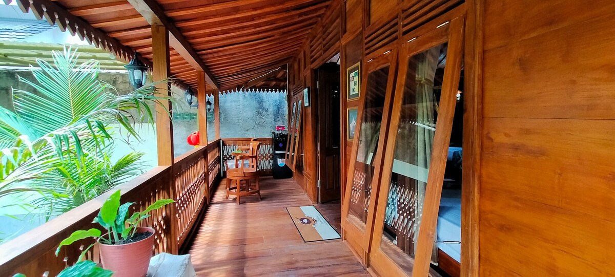 An inviting wooden terrace is displayed, featuring slatted roofs that provide shade. A dining table and chairs are arranged for outdoor enjoyment. Lush green plants are situated along the railing, contributing to the serene environment of the traditional wooden house.