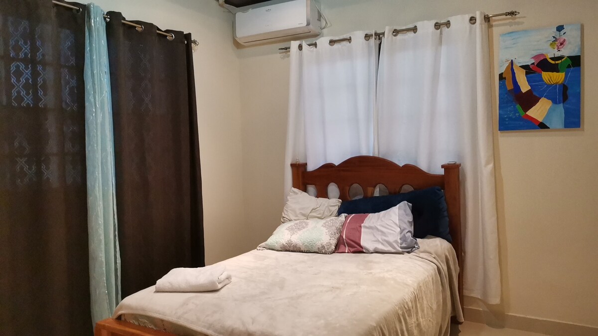 A comfortable sleeping area is presented with a spacious bed adorned with a light-colored blanket and decorative pillows. Natural light enters through sheer and opaque curtains, while a vibrant piece of wall art adds a pop of color to the neutral room.