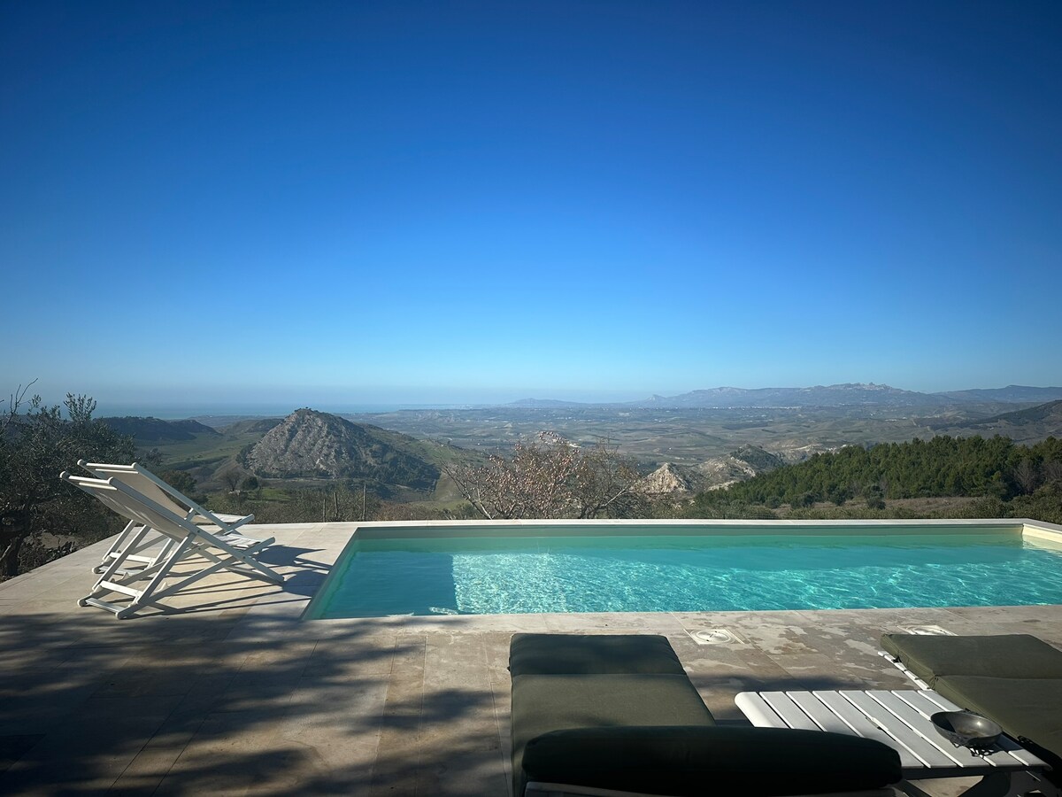 A serene pool area offers a clear view of the expansive valley and distant mountains. Lounge chairs are positioned by the water’s edge, inviting relaxation under a clear blue sky. The calm surface of the pool reflects the surrounding natural beauty.