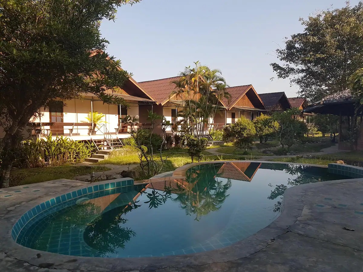 A serene pool reflects the surrounding greenery and wooden structures of the resort. Tropical plants border the water, creating a peaceful setting, while the morning light enhances the tranquil atmosphere of the outdoor space.