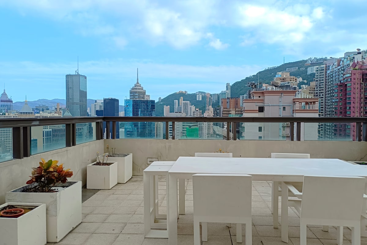 A rooftop terrace features a white dining table surrounded by matching chairs, providing an inviting space for outdoor meals. Planters with greenery frame the area, and panoramic city views are visible against a backdrop of distant mountains and towering skyscrapers.