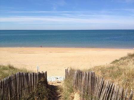 A sandy beach is seen from a pathway lined with wooden fencing, leading down to the calm ocean. The shoreline stretches into the distance under a clear blue sky, with gentle waves lapping at the sand.