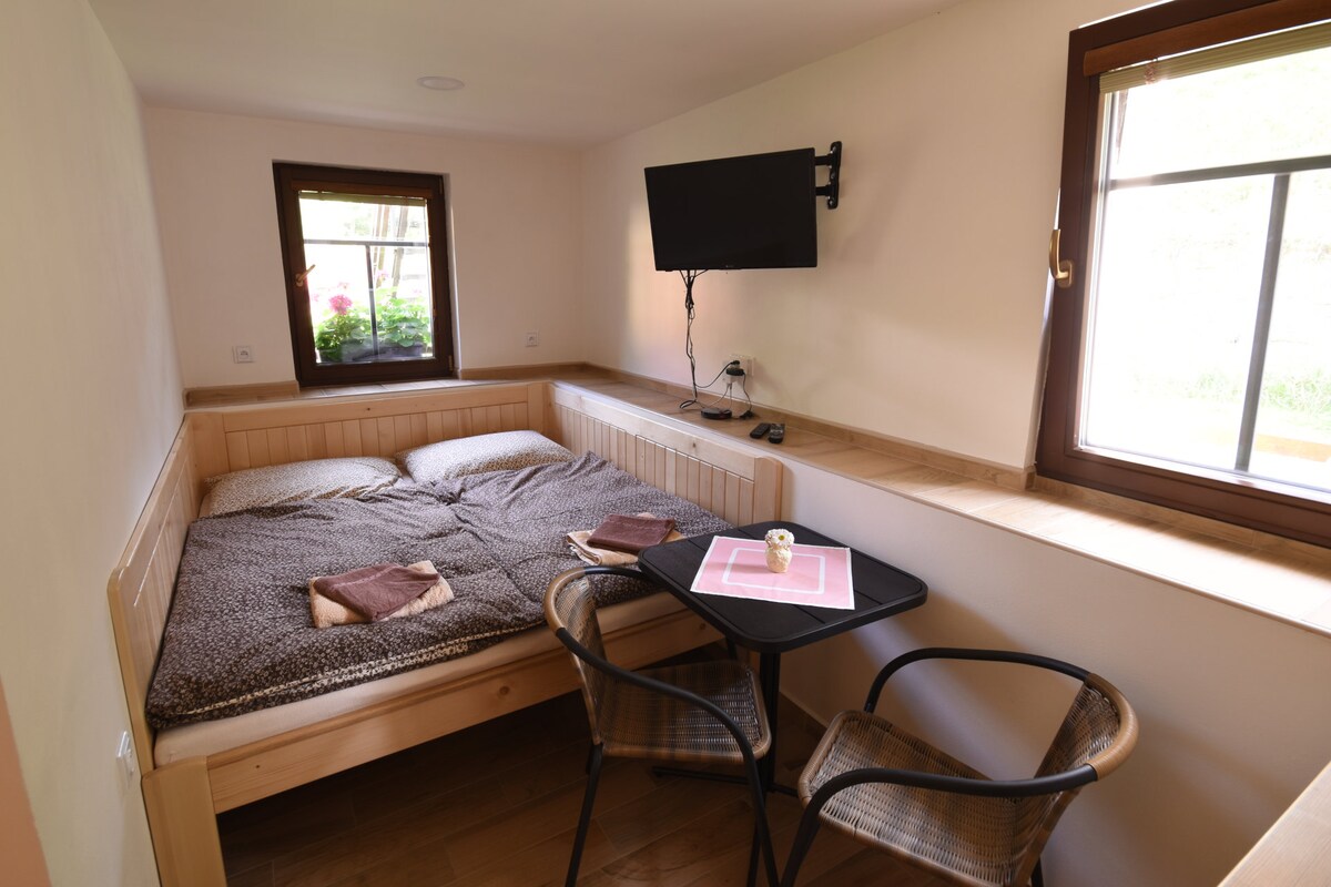 A compact sleeping area features a spacious bed with neutral bedding and two folded towels on top. A small dining table with two chairs is present, accompanied by a wall-mounted television. Natural light filters through the window, illuminating the warm wooden accents.