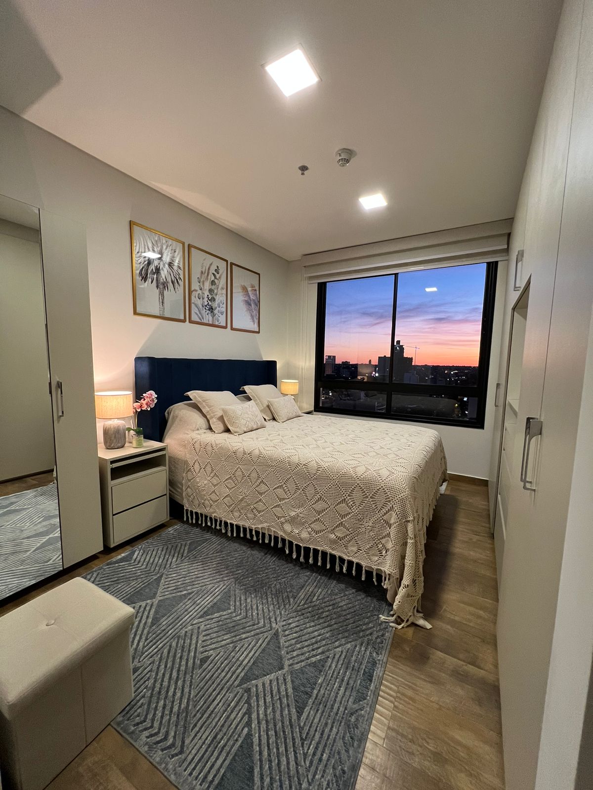 A serene bedroom is observed, featuring a spacious bed adorned with a light beige throw and three decorative pillows. A large window presents a city skyline during sunset, while subtle lighting illuminates the space. Mirrored furniture adds a touch of elegance to the room.