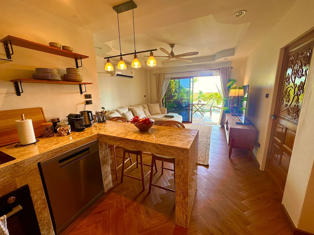A modern kitchen space features a light-colored stone countertop, equipped with a coffee machine and amenities. The open-plan layout connects to a living area with a luxurious sofa facing a large sliding door leading to a terrace with outdoor seating.